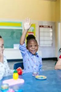 A young girl actively participates in a classroom, raising her hand to answer a question.